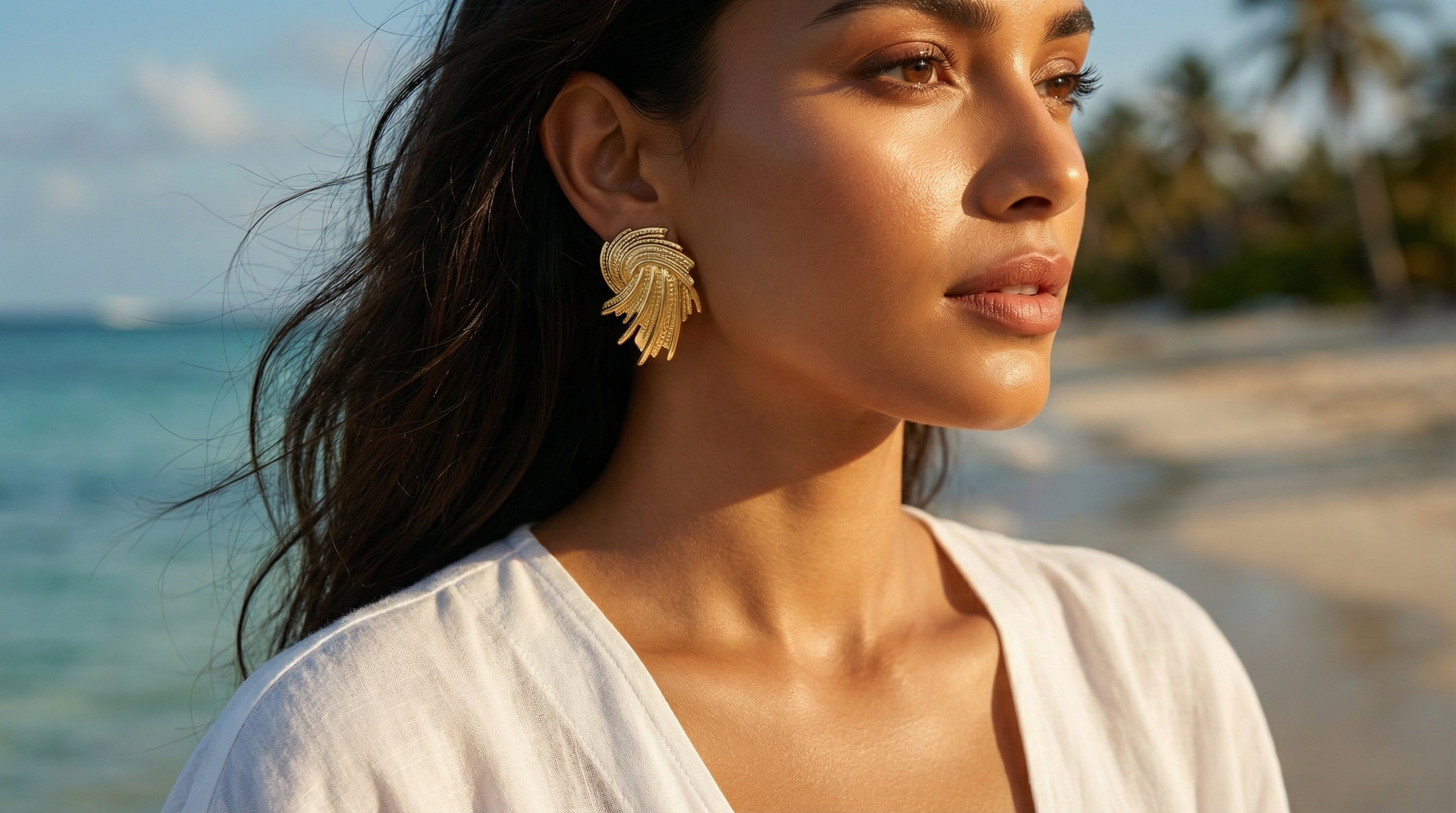Woman wearing gold earrings on a beach with ocean and palm trees in the background