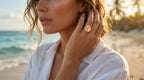 Woman on a beach wearing a white shirt and a large ring, with a blurred background of ocean and sky.