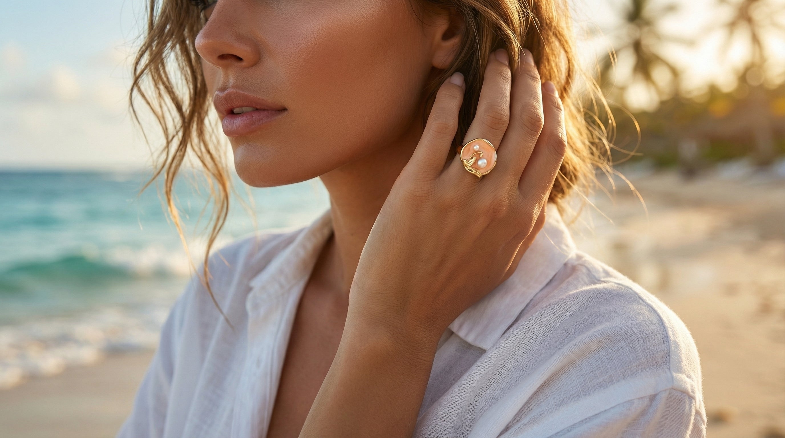 Woman on a beach wearing a white shirt and a large ring, with a blurred background of ocean and sky.