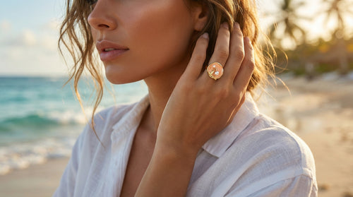 Woman on a beach wearing a white shirt and a large ring, with a blurred background of ocean and sky.
