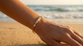 Hand wearing a gold bracelet on a beach with ocean in the background
