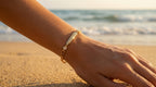 Hand wearing a gold bracelet on a beach with ocean in the background
