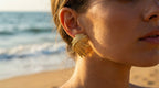 Close-up of a person wearing gold earrings with a beach in the background