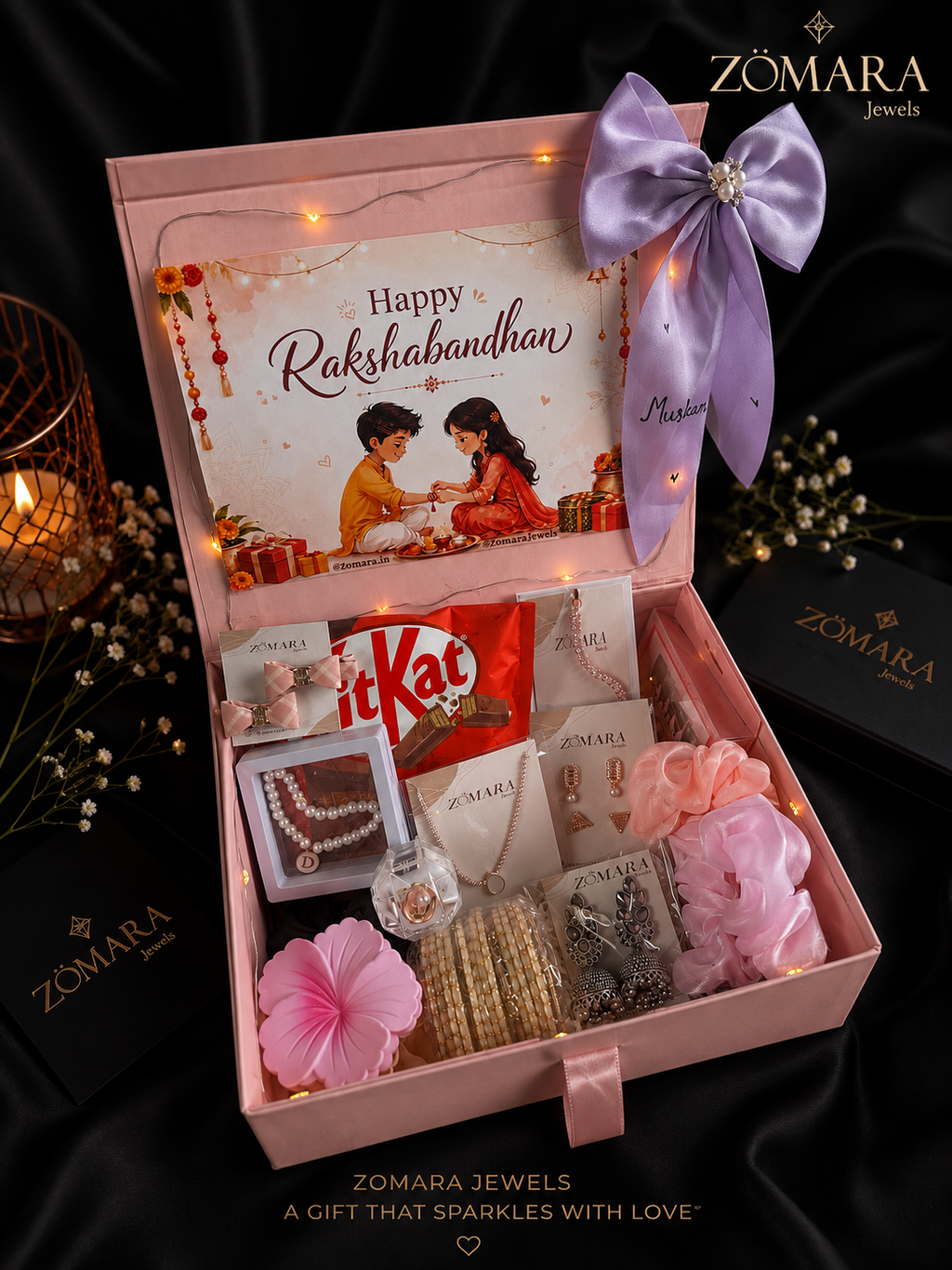 An open pink ZOMARA Jewels gift box arranged with a large lavender ribbon, warm fairy lights, jewelry pieces, a card reading Happy Rakshabandhan, a KitKat bar, and pink decorative flowers on a black satin background.