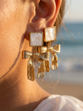 Gold bow-shaped earrings worn by a person with a blurred beach background