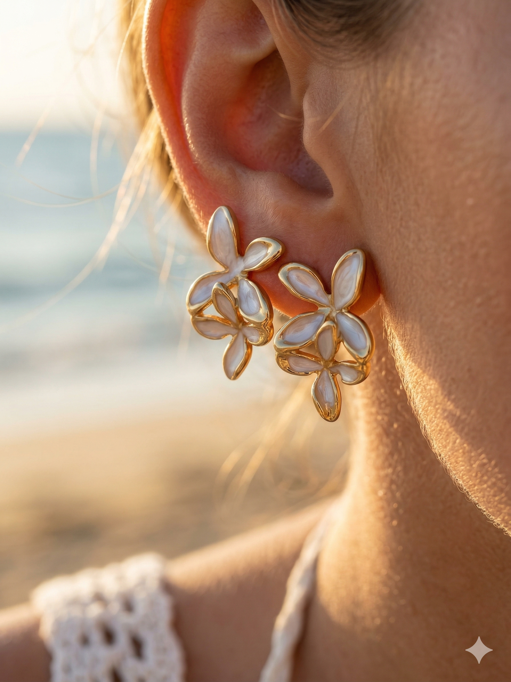 Close-up of a person wearing gold and white floral earrings with a blurred beach background.