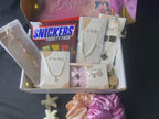 Gift box with jewelry, snacks, and decorative items on a bed with flowers and lights.