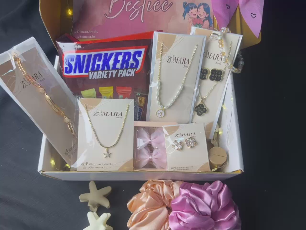 Gift box with jewelry, snacks, and decorative items on a bed with flowers and lights.