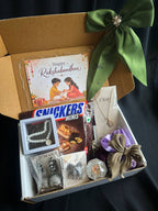 An open gift box containing multiple items: a Rakhi card, a Snickers Minis bar, a pearl necklace, silver earrings and jewelry pieces, a purple and gold hair bow, and a green satin ribbon on the box lid, photographed on a dark fabric background.