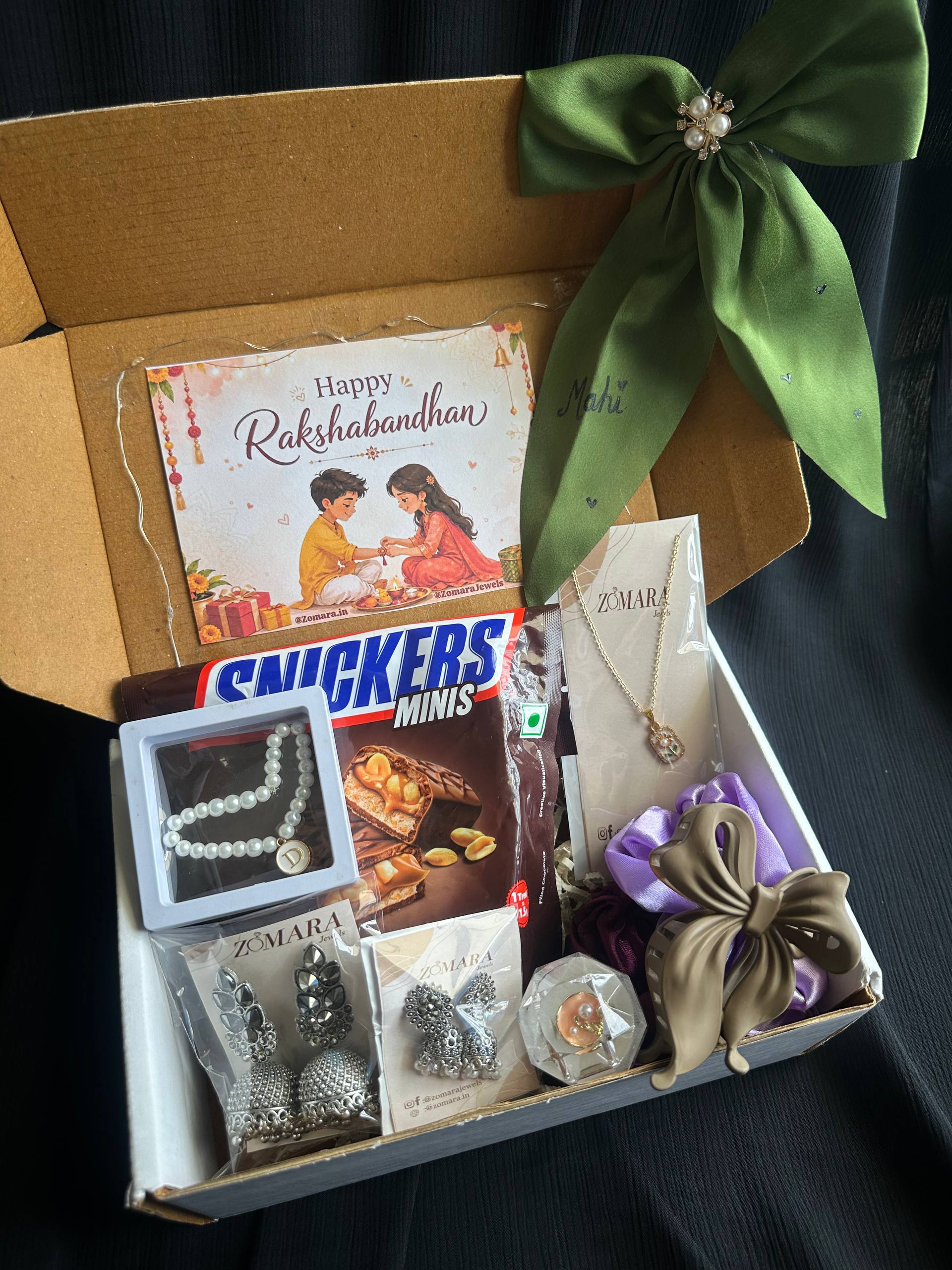 An open gift box containing multiple items: a Rakhi card, a Snickers Minis bar, a pearl necklace, silver earrings and jewelry pieces, a purple and gold hair bow, and a green satin ribbon on the box lid, photographed on a dark fabric background.