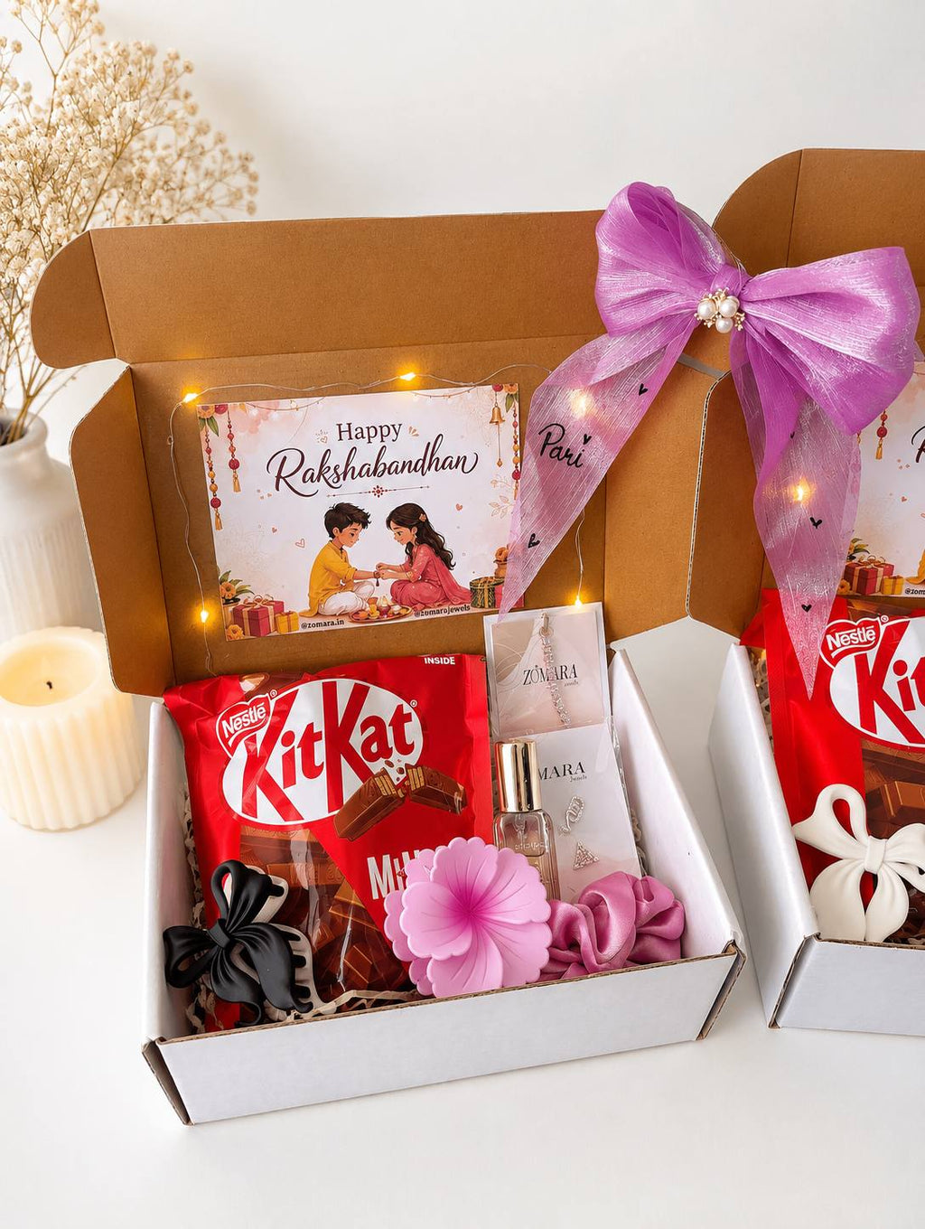 A Raksha Bandhan gift box set on a white surface, featuring a brown cardboard box filled with a pink Raksha card, a red bag of KitKat, small perfume bottles, a pink flower, and a black ribbon, tied with a large purple satin bow. Another partial box is visible on the right.