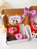 A Raksha Bandhan gift box set on a white surface, featuring a brown cardboard box filled with a pink Raksha card, a red bag of KitKat, small perfume bottles, a pink flower, and a black ribbon, tied with a large purple satin bow. Another partial box is visible on the right.