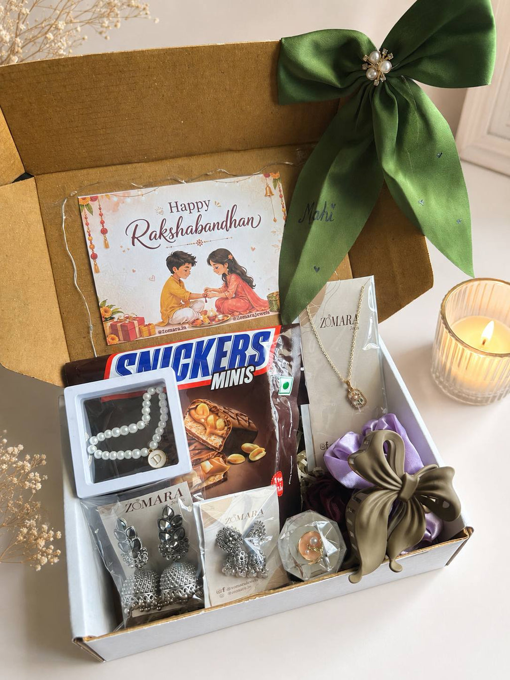 A curated gift box set on a light surface, featuring a brown cardboard box with a large green satin bow, a Rakhi card, Snickers Minis chocolate, silver jewelry, and other gift items. A lit candle and dried flowers are placed beside the box.