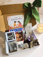 A curated gift box set on a light surface, featuring a brown cardboard box with a large green satin bow, a Rakhi card, Snickers Minis chocolate, silver jewelry, and other gift items. A lit candle and dried flowers are placed beside the box.