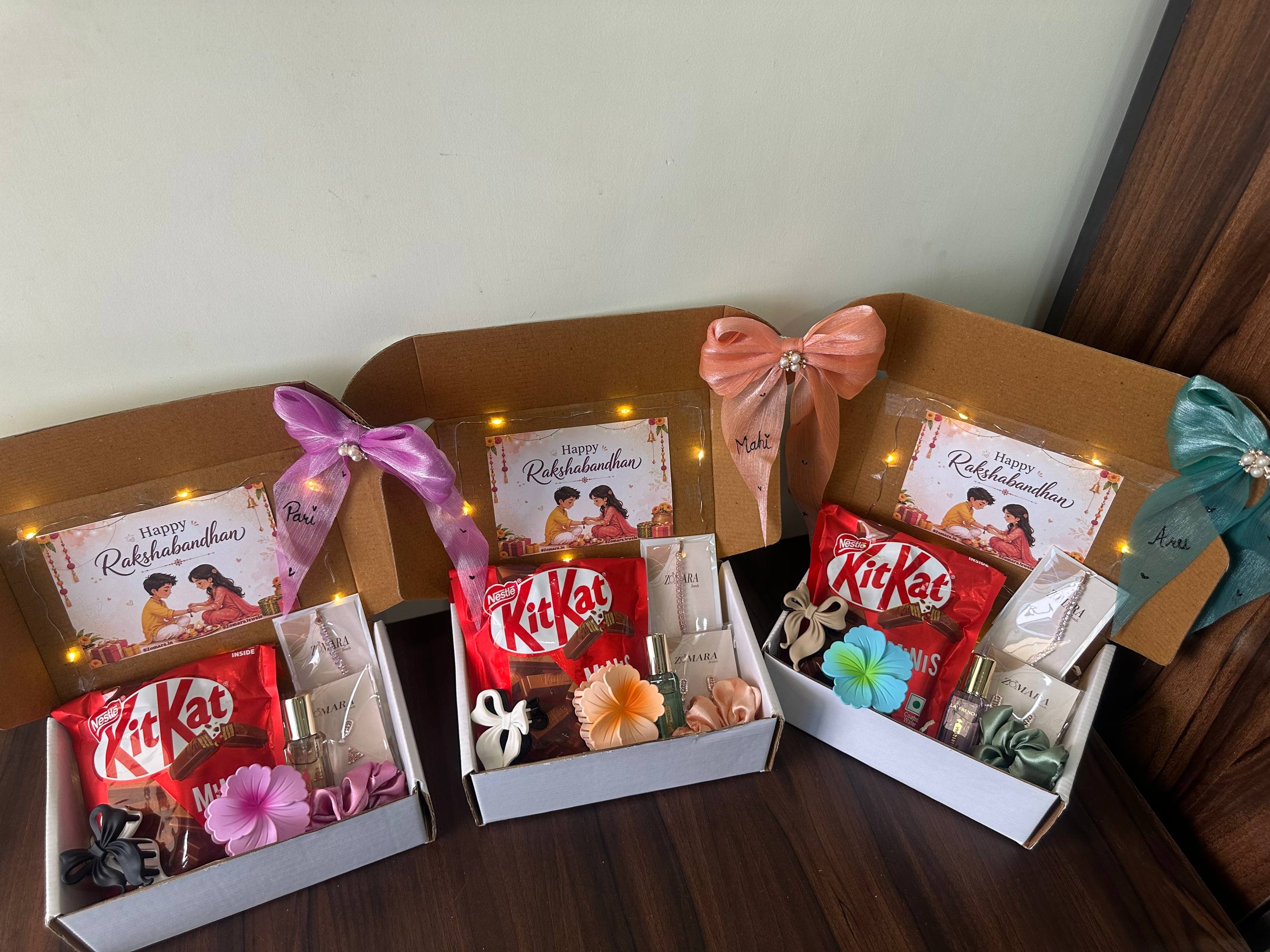 Three open gift boxes arranged on a dark wooden surface, each containing a red KitKat, a small card, decorative flowers, and ribbon bows. The boxes are photographed indoors against a plain light wall.